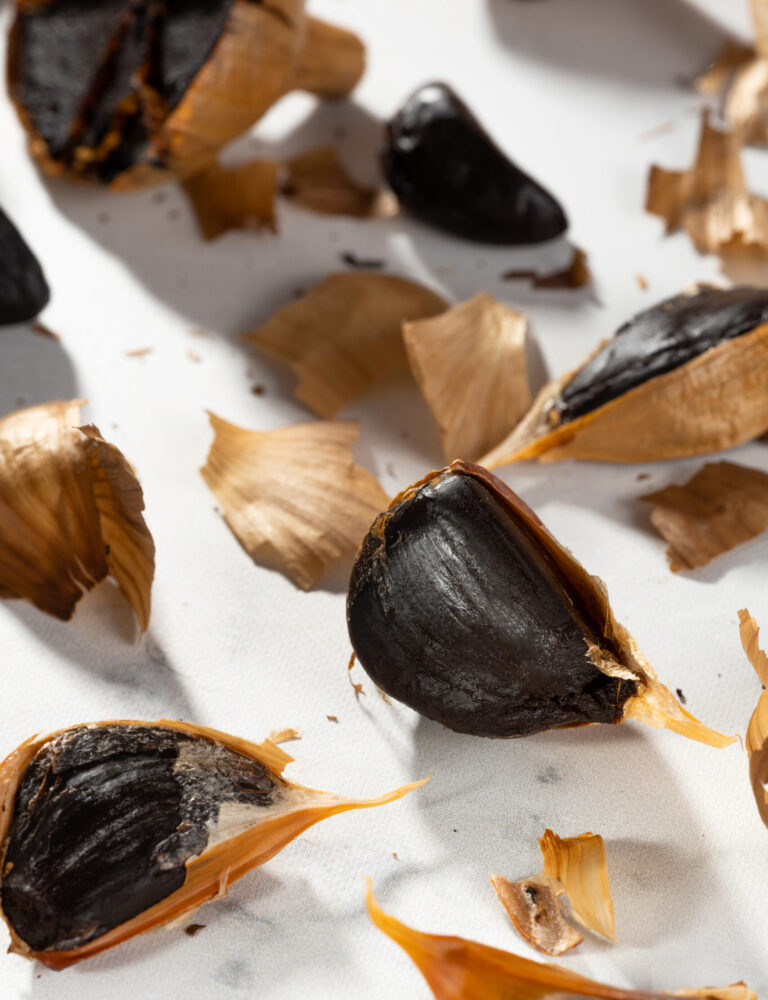 Cloves of black garlic lying on a white background. Mess on the kitchen table with peel and fermented garlic cloves. Fermented foods, healthy food, vegan gastronomy, self-care, close-up, hard shadow